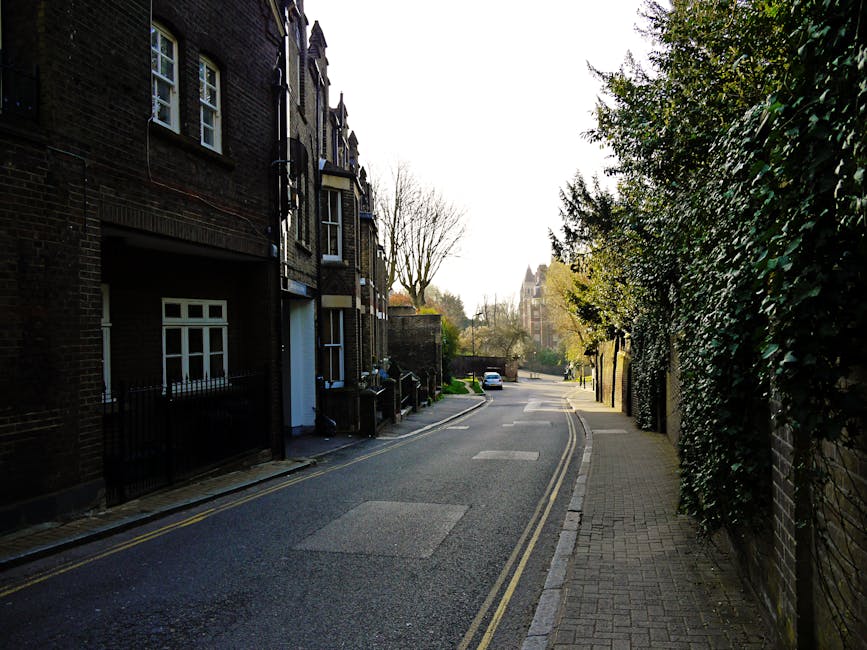 A quiet residential street in West Hampstead with a narrow pavement on the right side lined by a brick wall covered in dense green foliage, and a row of Victorian-style terraced houses on the left, featuring dark brick facades, white window frames, and small front gardens enclosed by black metal fences. Visible in the scene, a white van parked further down the street near the end of the visible pavement, suggesting vehicle access for home relocation or furniture transport. The street is illuminated by soft daylight, with leafless trees in the background indicating a late autumn or early winter setting. The scene depicts a typical urban environment suitable for house removals and professional moving services, emphasizing the accessibility of the area for loading or unloading furniture and moving boxes, with no visible action but implying a typical moving process or preparations.