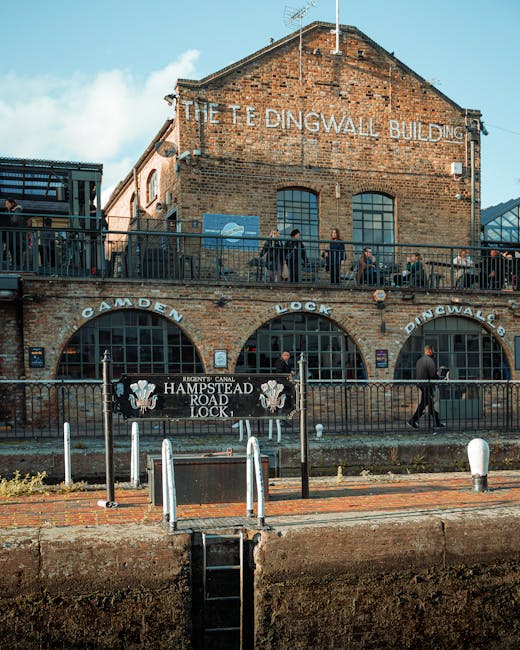 A multi-level brick building labeled 'The Dingwall Building' with large arched windows on the lower floor and smaller rectangular windows on the upper floor. A metal railing runs along the upper platform where several people are standing, some engaged in conversation. The lower level features a sign that reads 'Camden Lock' and 'Hampstead Road Lock,' with decorative elements and smaller signs indicating the lock's historical significance. In the foreground, there is a paved area with low white bollards and a short set of stairs leading down to a canal or waterway visible at the bottom of the image. The scene is illuminated with natural daylight, highlighting the industrial architecture typical of a historic canal lock location, relevant to transport and moving logistics, as seen in house removal services by companies like Man with Van West Hampstead.