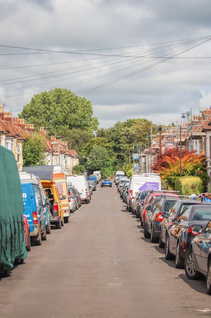 A busy street scene in West Hampstead featuring parked cars along the left side of the paved roadway, with several residential and commercial buildings displaying colorful facades and storefronts. Ped