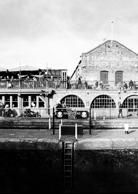 A black and white photograph of The Tea Dingwall Building, a brick structure with large arched windows on the ground floor and smaller rectangular windows above, situated next to a waterfront. In front of the building, there is a metal railing and a sign reading 'Hampstead Road Lock,' with a small ladder descending from the pavement to the water's edge. The scene shows a busy outdoor area with tables, chairs, and umbrellas on a terrace above the waterfront, suggesting a dining or social space, with pedestrians walking along the street. The sky appears partly cloudy, and the scene is well-lit, capturing the historic architecture and lively atmosphere typical of a central urban location in West Hampstead. This setting is relevant for house removals or relocation services, illustrating a typical urban environment where furniture transport and home moving logistics may occur, with glimpses of the transport infrastructure seen in the background.