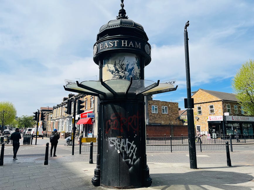 A black, round information kiosk with a domed top and a glass display section positioned on the pavement at the entrance to East Hampstead, with a large sign reading 'EAST HAM' on the upper part. The kiosk is surrounded by metal fencing and is situated near a busy street intersection, featuring several pedestrians walking nearby. Behind it, there are multi-story brick buildings with shopfronts, including a nail salon, and other commercial premises. The sky above is partly cloudy with patches of blue, and a few green-leaved trees are visible on the right side of the image. This urban scene captures the regular setting where house removals and furniture transport services by Man with Van West Hampstead may operate, with the area suitable for loading and moving activities near local shops and residential areas.