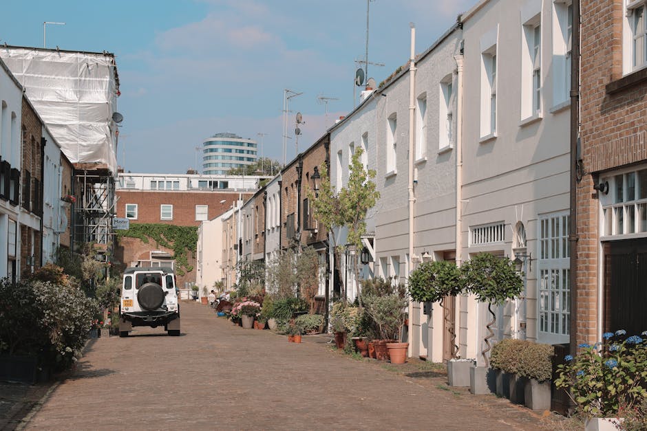 A view of a narrow residential street in West Hampstead, NW6, with a white van parked on the cobblestone pavement adjacent to row of terraced houses. The houses have white and brick exteriors, with some featuring small front gardens with potted plants and greenery. The van, used for home relocation and furniture transport, is positioned near the entrance of one property, suggesting an ongoing loading process. Resting against the van, there is a person’s hand visible, possibly involved in the packing or unloading activities, with cardboard boxes and plastic wrapping materials nearby. The scene is well-lit with natural daylight, showing a clear sky and modern buildings in the background, indicating an urban setting suited for professional removals and packing services, such as those provided by Man with Van West Hampstead.