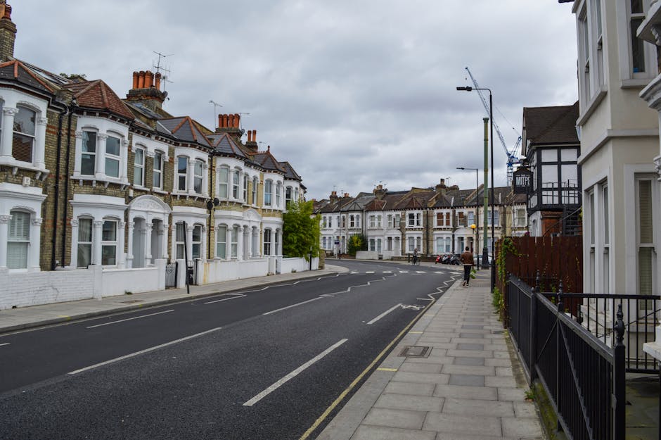 A street in West Hampstead featuring a row of Victorian-style terraced houses with white facades and red-tiled roofs, some with bay windows and chimneys. The pavement runs along the right side of the image, with black metal railings in front of a white residential building. A few pedestrians are visible walking along the sidewalk. Overcast skies with grey clouds cast diffused light over the scene. In the background, there are construction cranes indicating ongoing development. The scene depicts a typical residential area suitable for house removals, with the street prepared for vehicle access to facilitate the loading process for a home relocation, with no moving boxes or furniture visible but an environment consistent with moving logistics managed by companies like Man with Van West Hampstead.