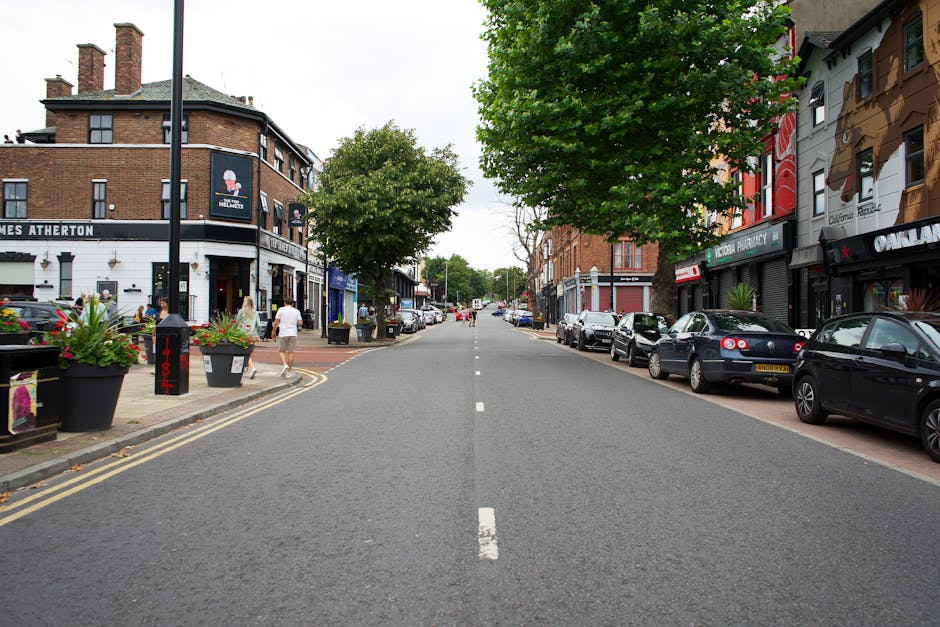 A straight view of a residential street in West Hampstead, with parked cars lining both sides of the road. On the left, there are pedestrians walking past shops, including a pub with a sign reading 'James Atherton', and large planters with flowers placed on the sidewalk. The buildings are multi-storey, with brick and painted facades, and some shop windows visible. On the right, more parked vehicles and storefronts are seen, with one building featuring a mural depicting a stylized animal face. The street is partly shaded by trees with green foliage, and the road surface is asphalt with a central white dashed line. The scene captures a calm moment in a busy neighbourhood, possibly during a home relocation process involving furniture and packing items being moved along this street, supported subtly by the presence of a removals service such as Man with Van West Hampstead.