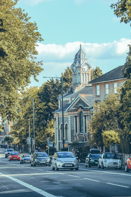 A street scene in West Hampstead featuring a line of parked cars along the roadside, with a historic church building visible in the background that has a clock tower topped by a pointed spire. The church's facade includes arched windows and decorative brickwork, partially obscured by surrounding deciduous trees with lush green foliage. The street is paved with asphalt and has white road markings, with traffic lights and street lamps along the pavement. In the context of a home relocation or furniture transport, the image depicts the typical environment where loading and unloading of removal vehicles may occur, as seen from a van or truck arriving or parked nearby, associated with professional removals services like Man with Van West Hampstead, supporting efficient packing and moving operations in the area.